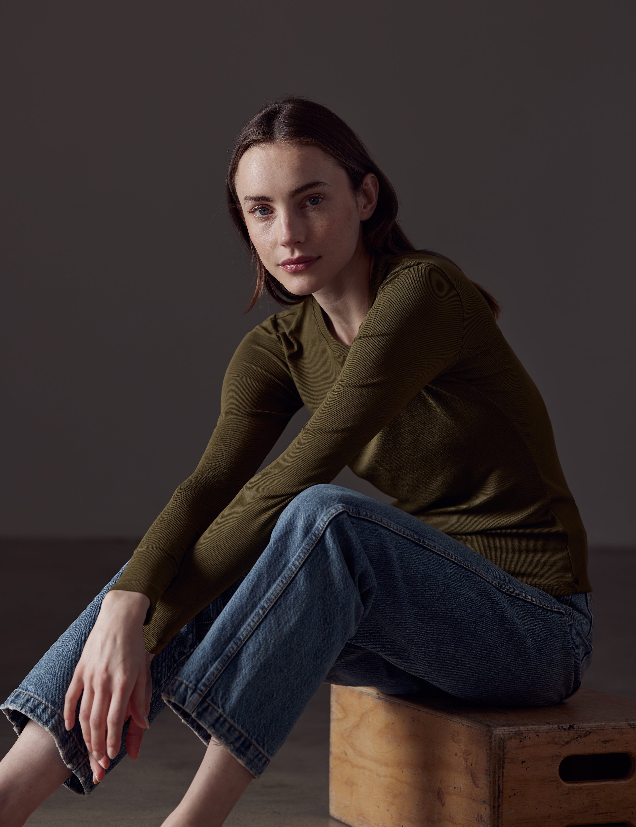 Front angled view of woman sitting on box wearing Legacy Long-Sleeve Crew in Command Green from AETHER Apparel.